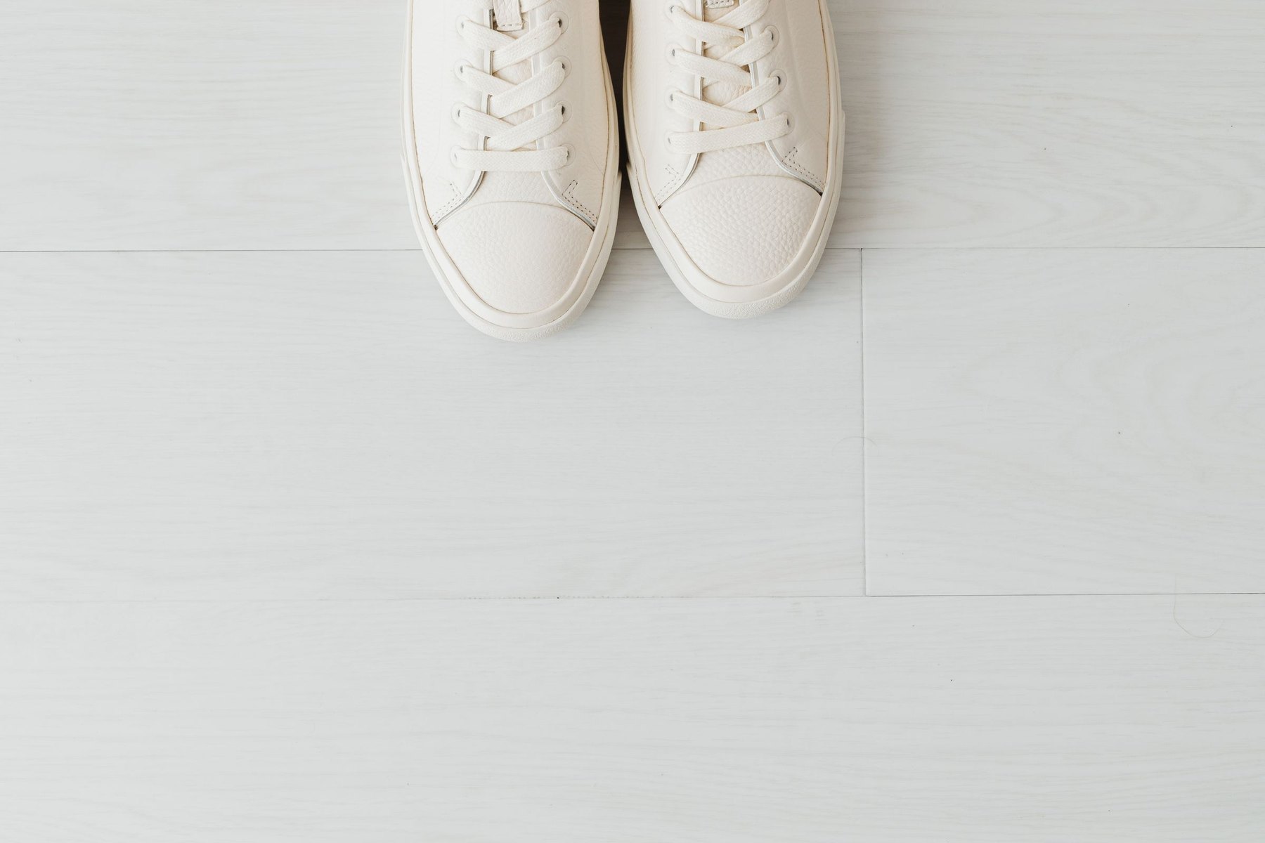 A pair of white full-grain leather low-top sneakers on a white wood floor.
